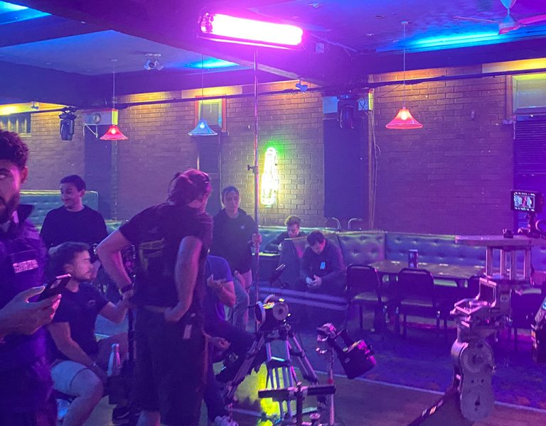 Several people are gathered in a neon-lit room with brick walls and coloured lights, some standing and some sitting on sofas. Camera equipment is visible in the foreground, suggesting a film or video shoot.