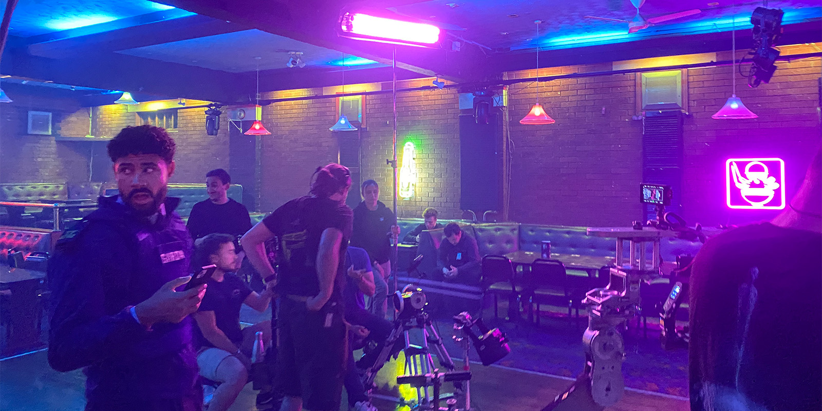 Several people are gathered in a neon-lit room with brick walls and coloured lights, some standing and some sitting on sofas. Camera equipment is visible in the foreground, suggesting a film or video shoot.
