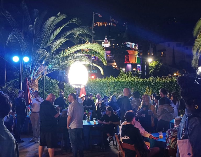 A large group of people gather outdoors at night under palm trees, illuminated by bright lights. Some are seated at tables whilst others stand and talk. Buildings and neon signs are visible in the background.