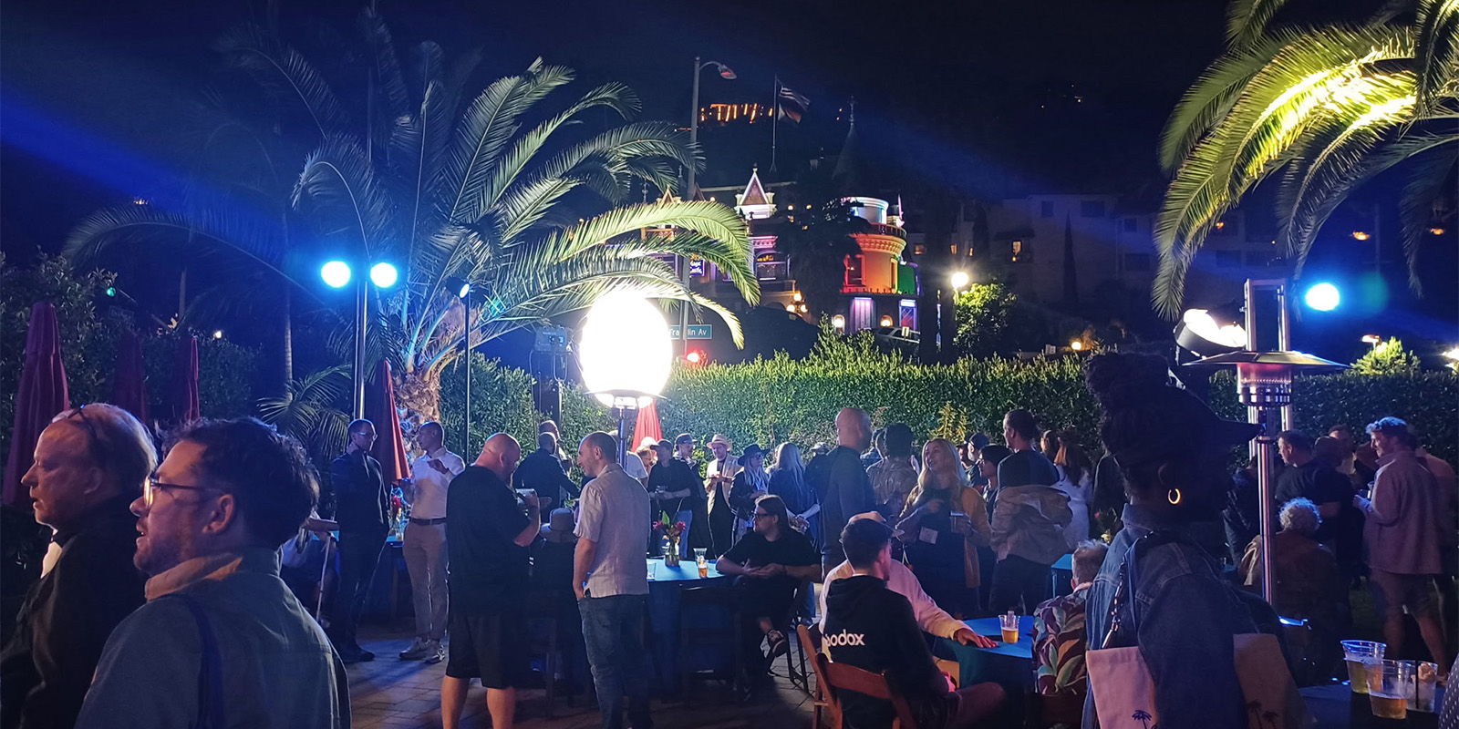 A large group of people gather outdoors at night under palm trees, illuminated by bright lights. Some are seated at tables whilst others stand and talk. Buildings and neon signs are visible in the background.