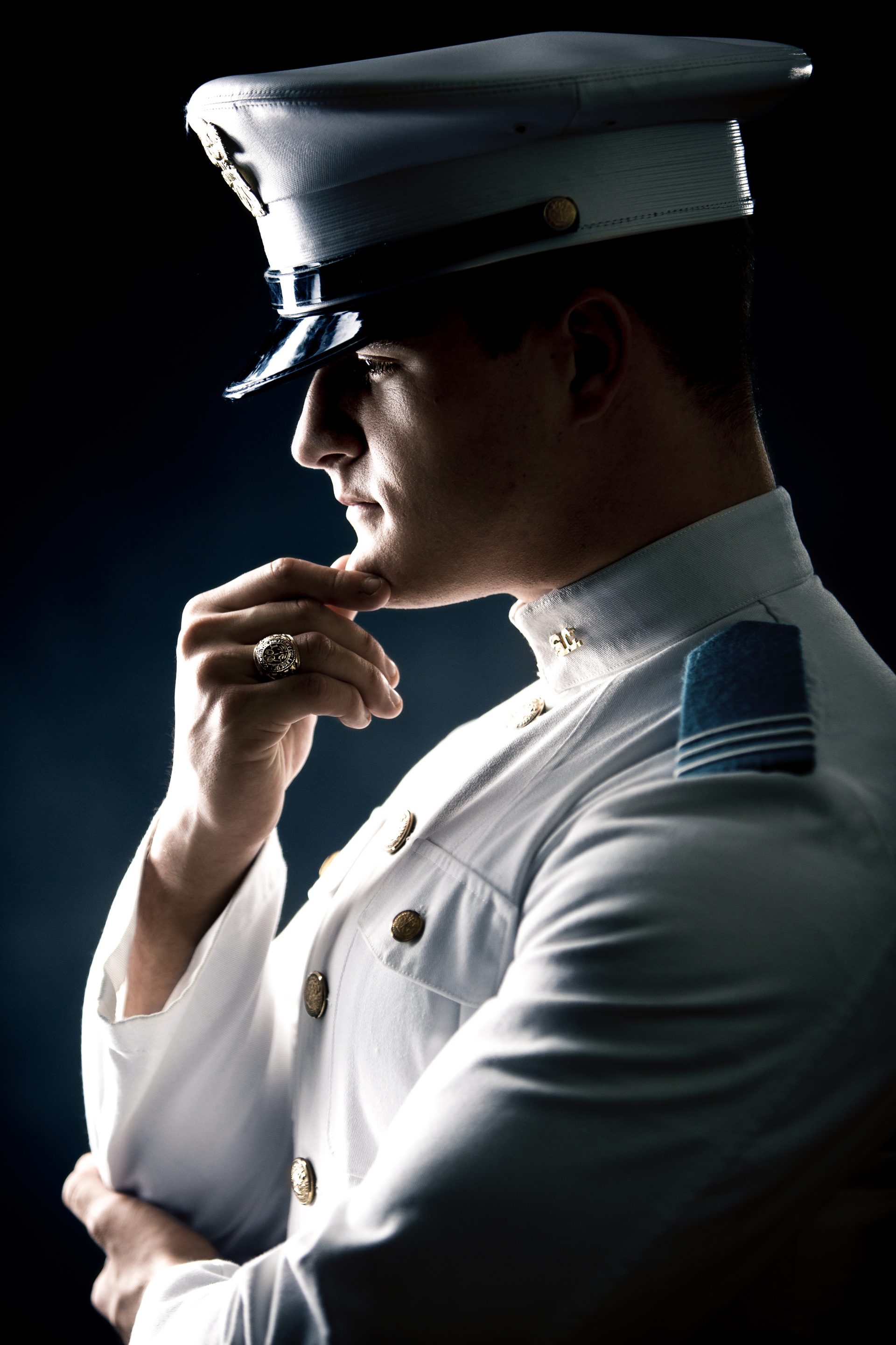 A man in profile wearing a white military dress uniform and hat stands against a dark background, resting his chin on his hand in a thoughtful pose.
