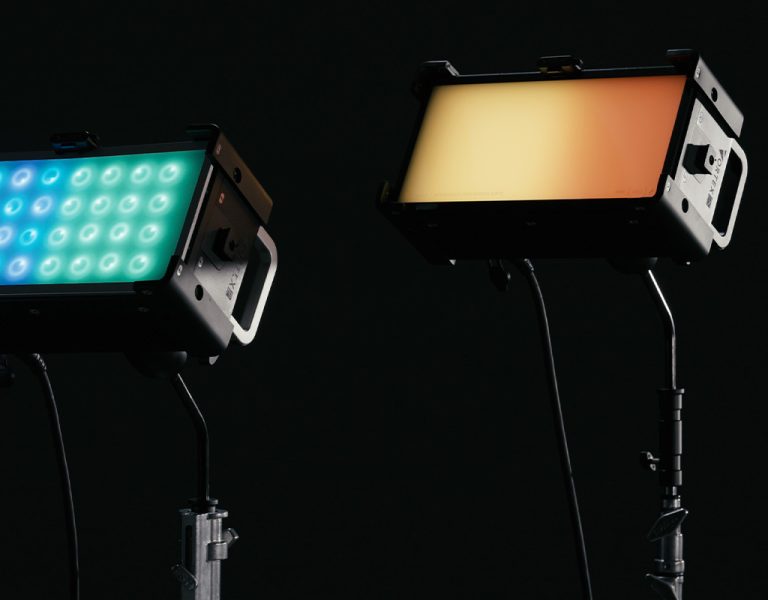 Two rectangular studio lights on stands against a black background; the left light displays multiple green and blue LED bulbs, whilst the right light emits a solid warm yellow-orange glow.