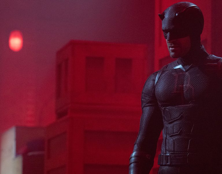 A person in a black superhero costume with small horns on the mask stands in a dimly lit room with red light and wooden crates in the background.