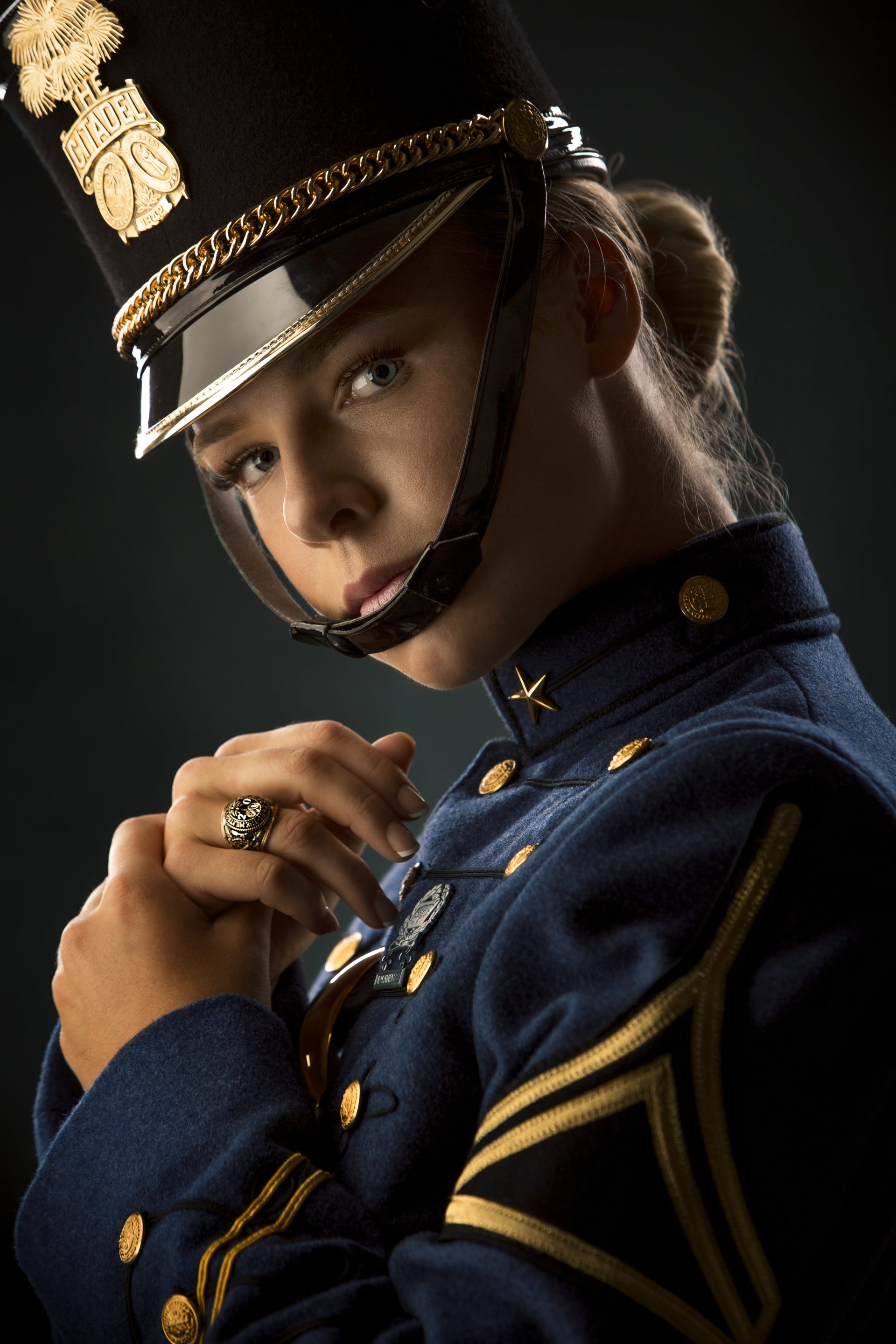A person wearing a formal dark blue military-style uniform and a tall hat with insignia, looking at the camera. The person holds their hands near their collar and wears a ring. The background is dark.