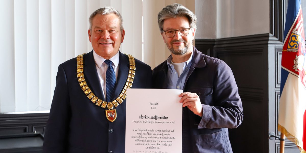 Two men stand indoors; one wears a gold ceremonial chain and suit, the other wears glasses and holds a large certificate. Both are facing the camera and smiling. A flag is partially visible in the background.