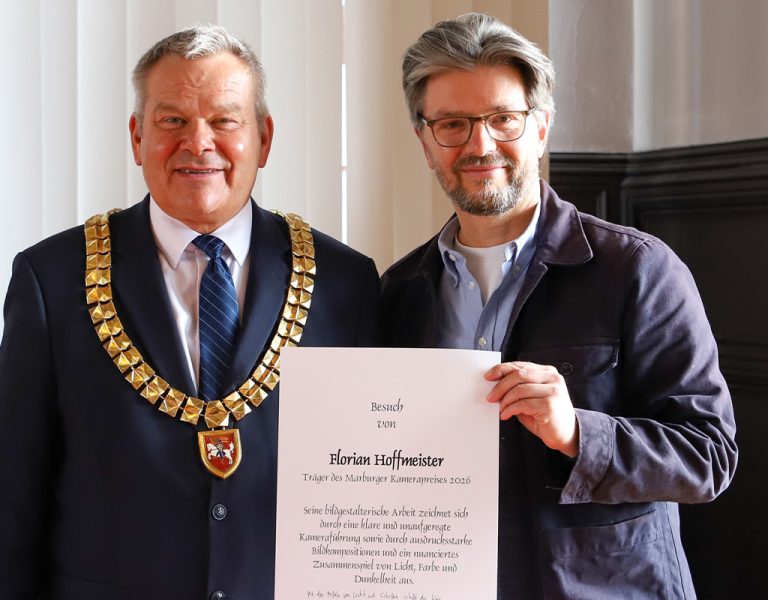 Two men stand indoors; one wears a gold ceremonial chain and suit, the other wears glasses and holds a large certificate. Both are facing the camera and smiling. A flag is partially visible in the background.