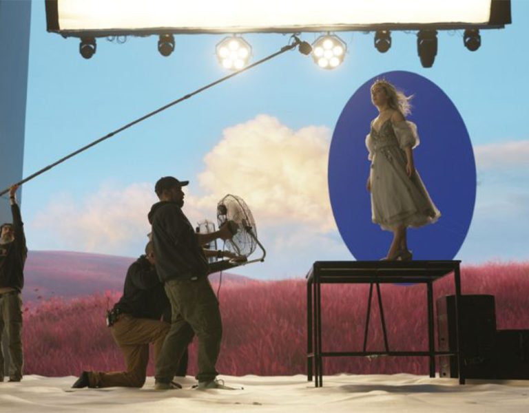 A film crew records a woman in a light dress standing on a platform in front of a large screen displaying a pink field and blue oval. Crew members hold equipment including a boom mic and a fan.
