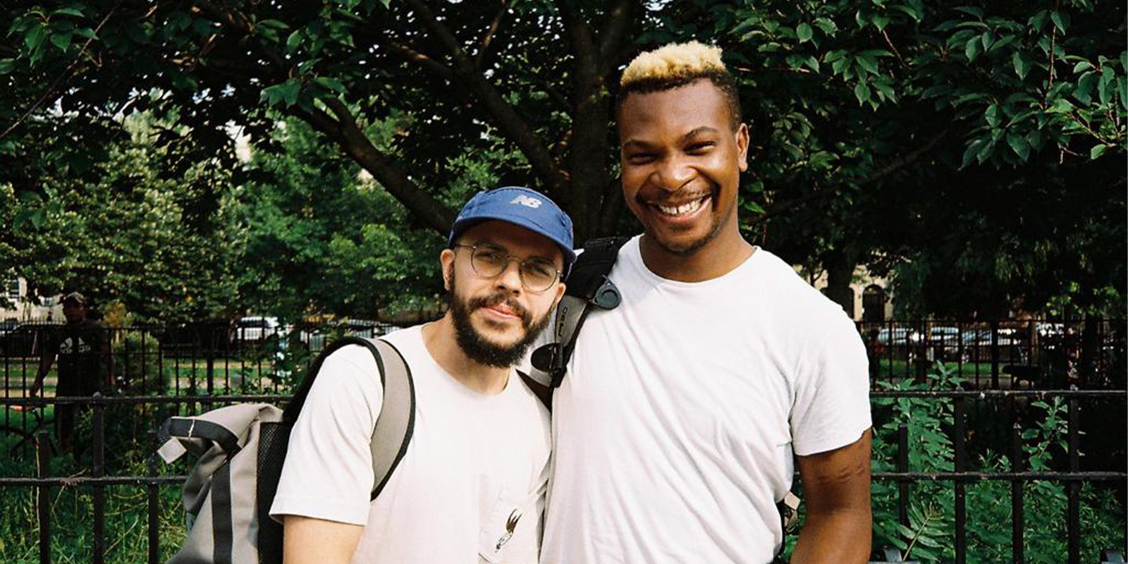 Two people stand close together outdoors in front of a black iron fence and green trees, both smiling. One wears a blue cap and glasses, the other has short blond hair. Both are dressed in white T-shirts and carry rucksacks.