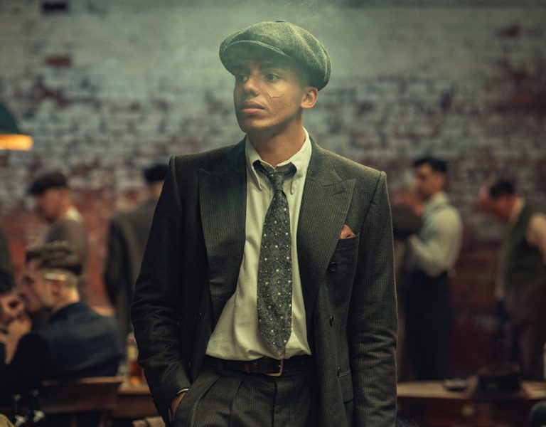 A man in a flat cap and suit stands in a dimly lit room with several other men seated at tables in the background, creating a vintage or historical atmosphere.