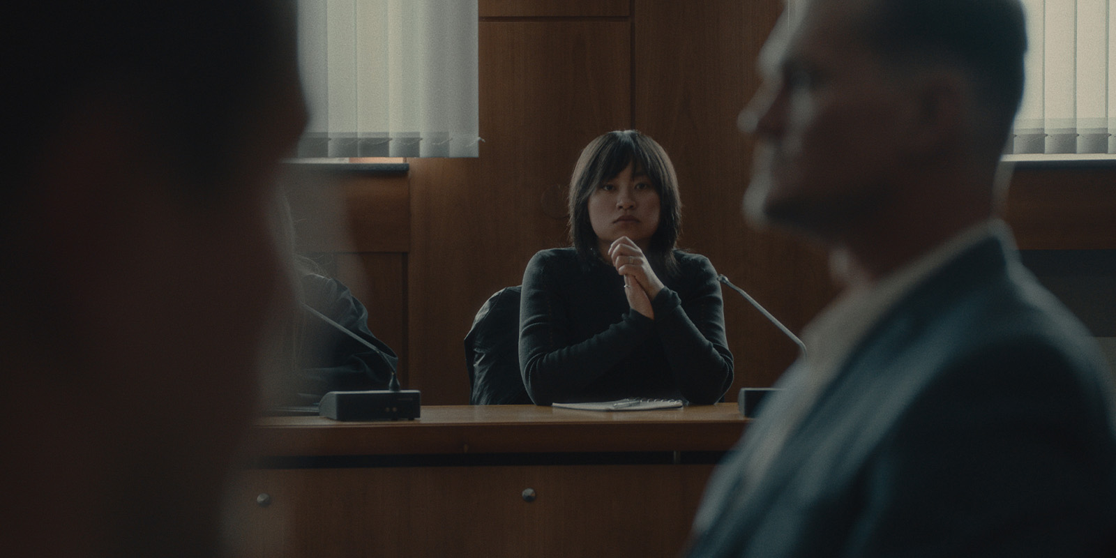 A woman sits at a table in a courtroom, looking forward with her hands clasped under her chin. Two people in the foreground are out of focus. The room has wood panelling and large windows with vertical blinds.