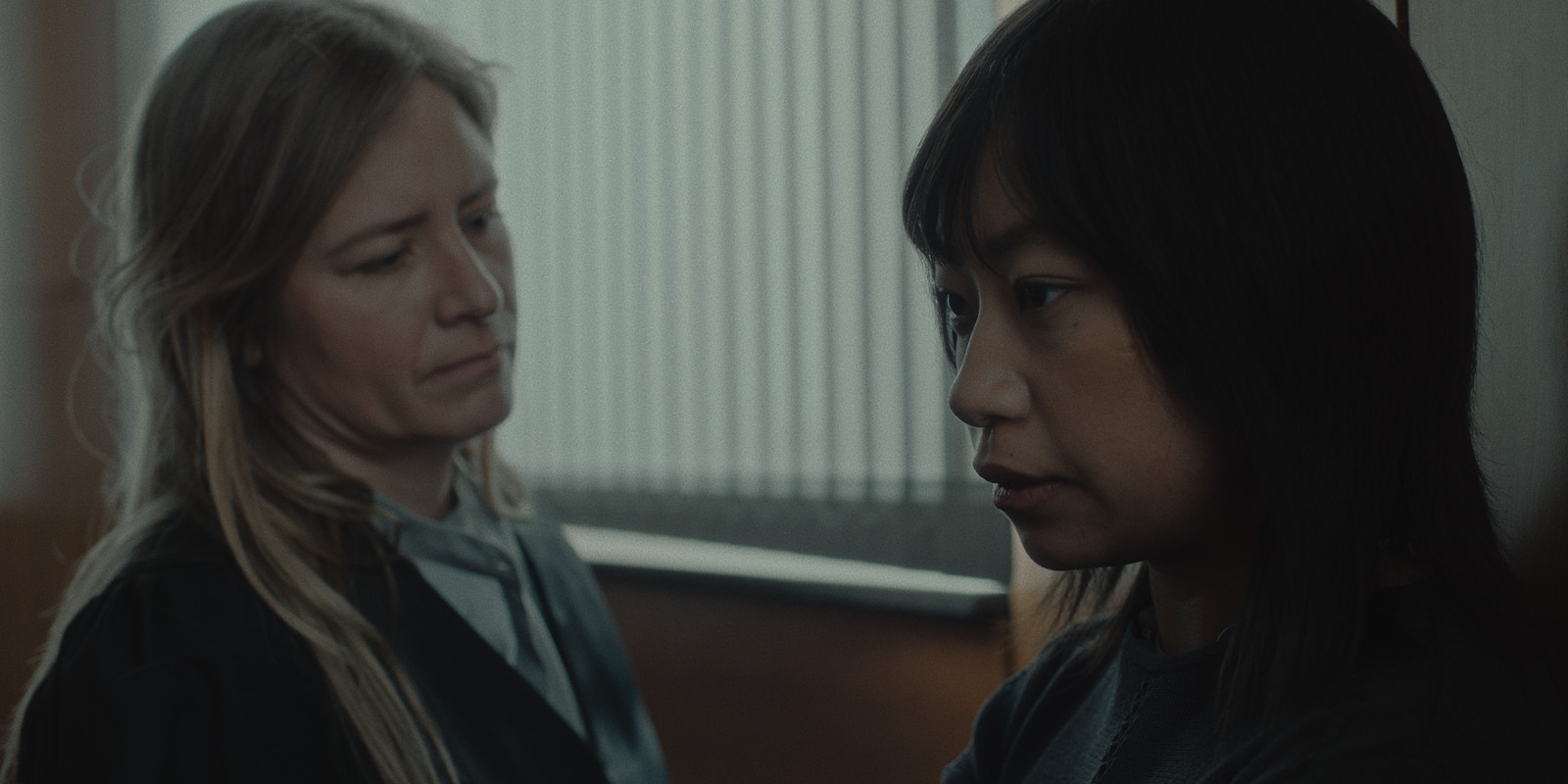 Two women stand indoors near a window with vertical blinds. One woman with long blonde hair looks at the other, who has dark hair and is facing away, appearing thoughtful or concerned. Both have neutral expressions.