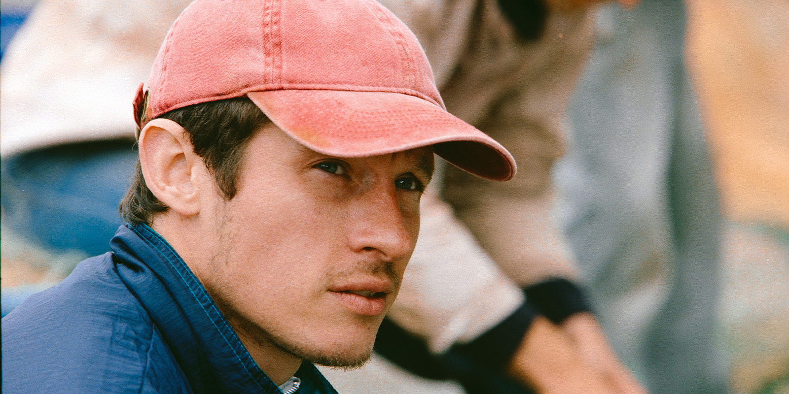 A man with light facial hair wears a faded red baseball cap and a blue jacket. He is looking slightly upwards. Another person is partially visible in the blurred background.