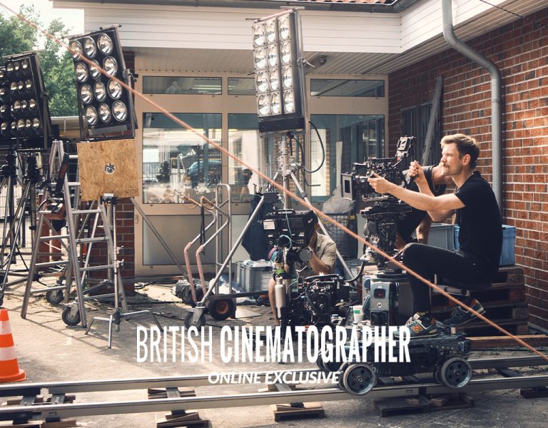 A man operates a camera on a dolly track surrounded by lighting equipment and cables outside a brick building. The text “BRITISH CINEMATOGRAPHER ONLINE EXCLUSIVE” appears at the bottom of the image.
