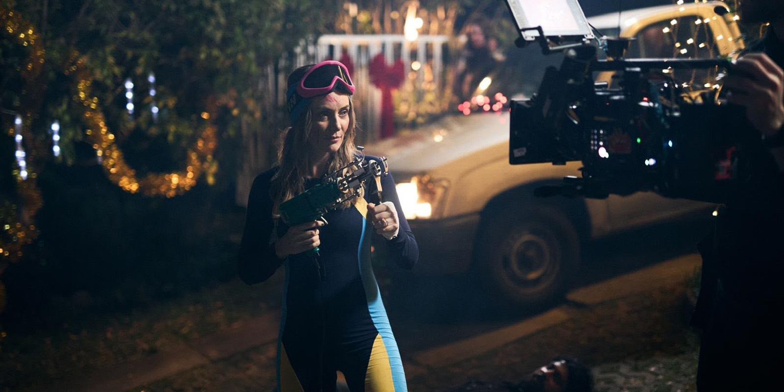A person in a wetsuit and pink goggles stands outdoors at night, holding a power drill. There are holiday lights and decorations in the background, a white vehicle, and a large camera filming the scene.