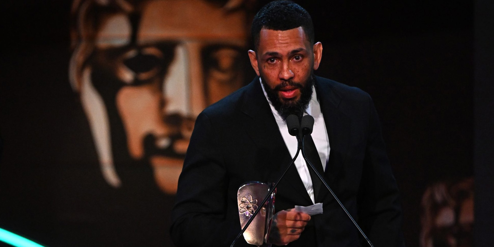 A man in a black suit stands at a microphone holding a BAFTA award and a piece of paper. A large BAFTA mask logo is visible in the background.