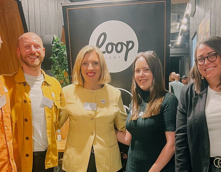 Six people stand indoors in front of a sign that reads Loop Talent. They are smiling at the camera and wearing name badges. The setting appears to be an event or gathering.