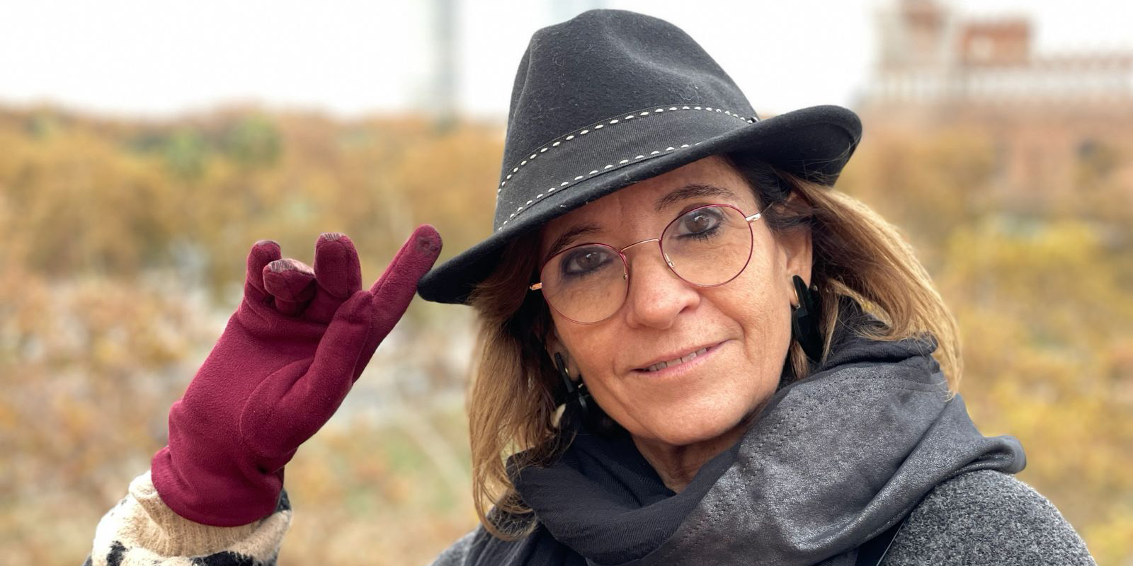 A woman wearing glasses, a black hat, a dark scarf, and a maroon glove is outdoors. She is touching the brim of her hat and looking at the camera. The background is blurred with autumn colours.