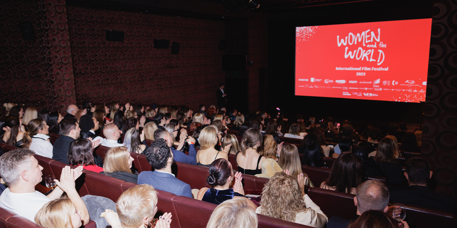 A full audience sits in a dark theatre watching a presentation on a large screen reading “WOMEN and the WORLD International Film Festival 2025.” The crowd is facing forward, and a speaker stands near the screen.