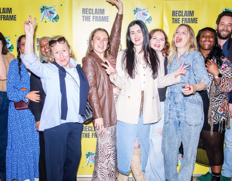 A group of twelve people stand together in front of yellow Reclaim the Frame banners, smiling and posing for the camera, with several making peace signs or raising their arms.