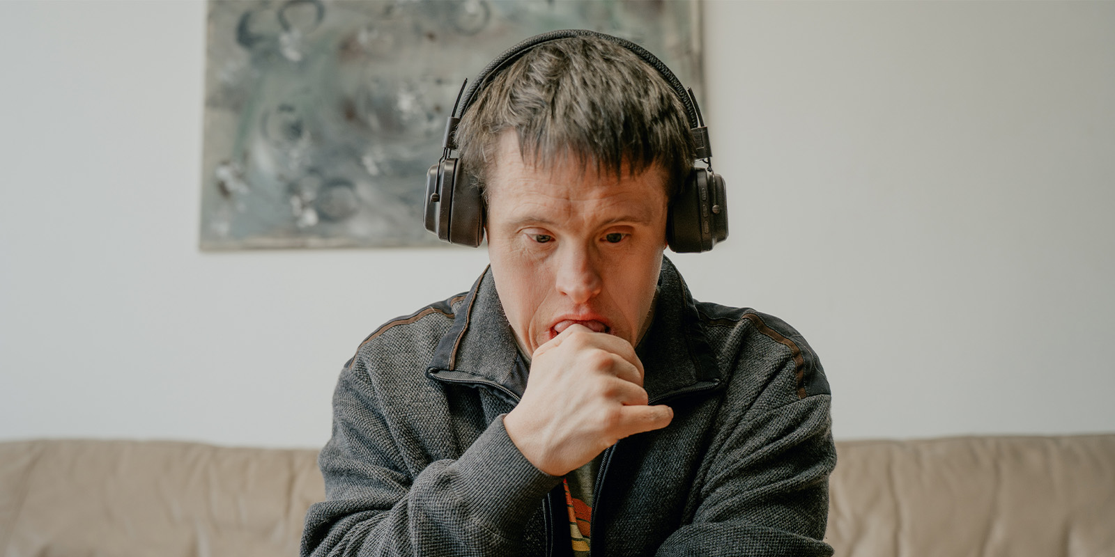 A man with Down’s syndrome sits on a sofa, wearing headphones and a grey jacket, looking down with his hand touching his mouth. A painting hangs on the wall behind him.