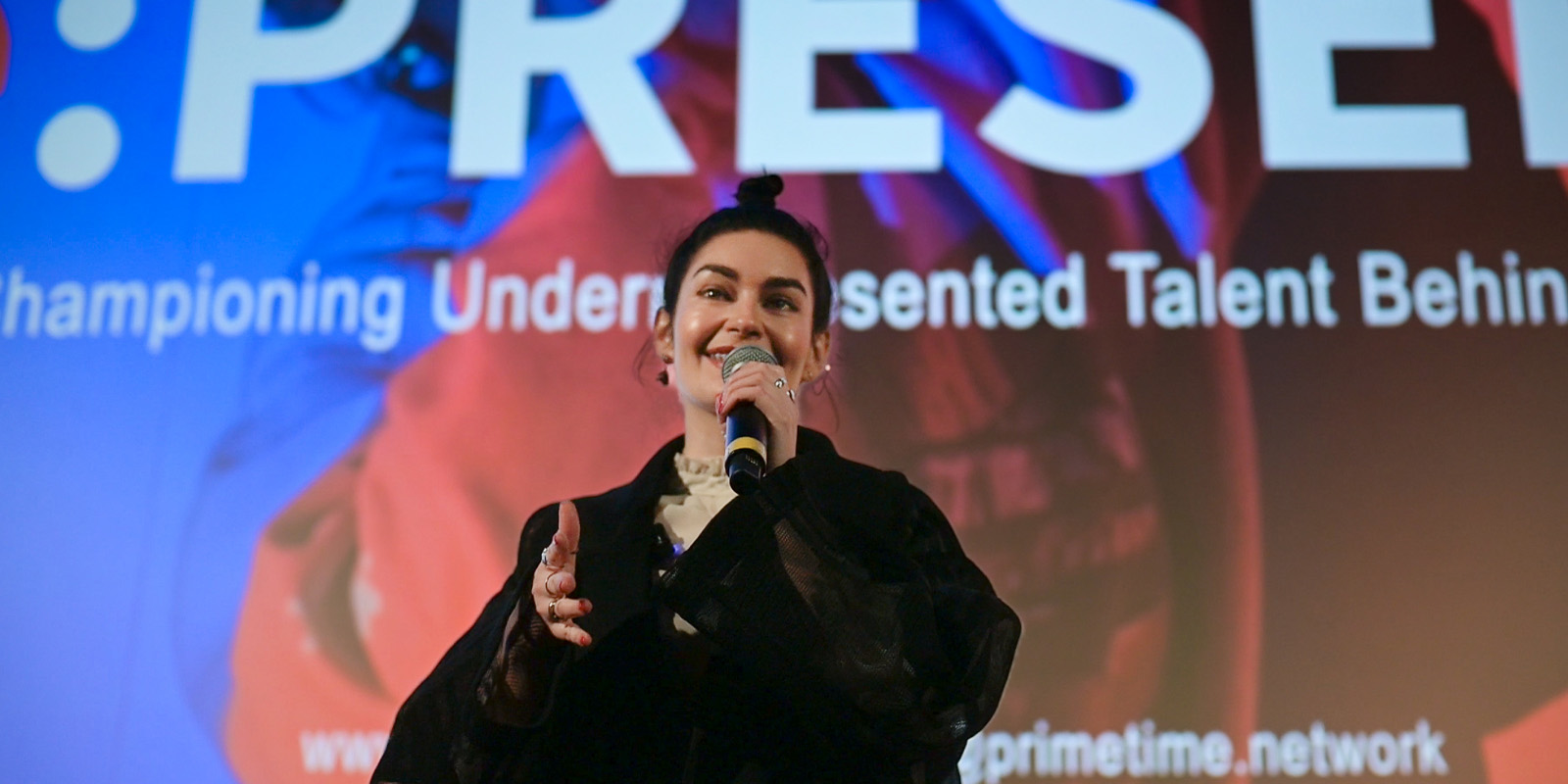 A person holding a microphone speaks on stage in front of a large screen that displays text about championing underrepresented talent behind the scenes.