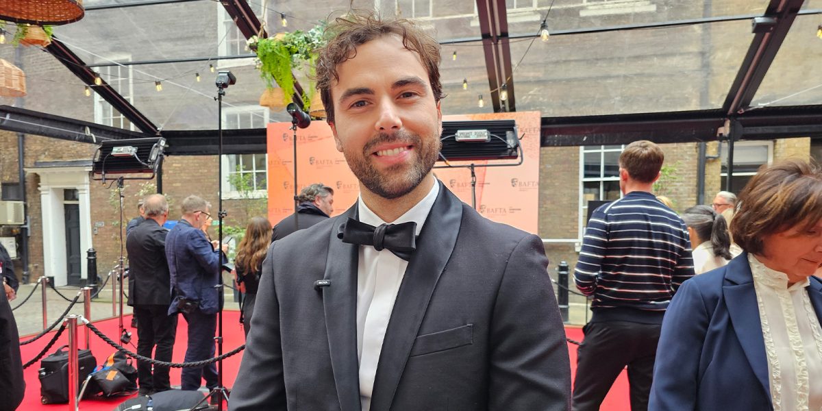 A man in a black dinner jacket stands smiling on a red carpet at an outdoor event, with several people, cameras, and a roped-off area visible in the background.