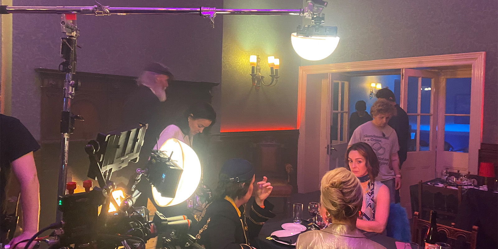 Several people are on a film set; two women sit at a dining table with cameras and lights focused on them, while crew members move around in the background. The walls are dimly lit and props are visible in the room.