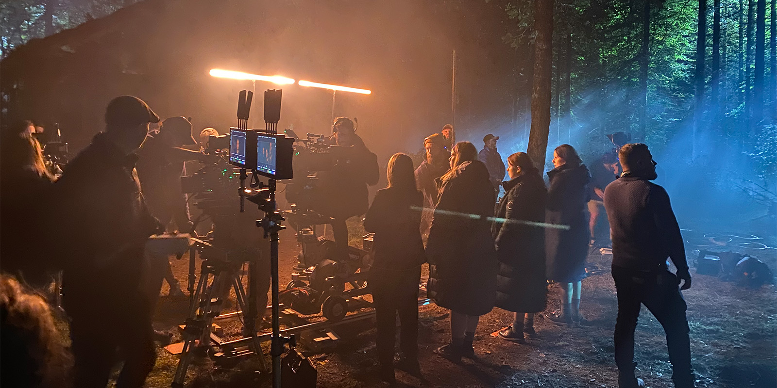 A night-time film set in a forest shows crew members operating cameras and equipment, with bright lights illuminating actors and crew standing together among tall trees and light mist.