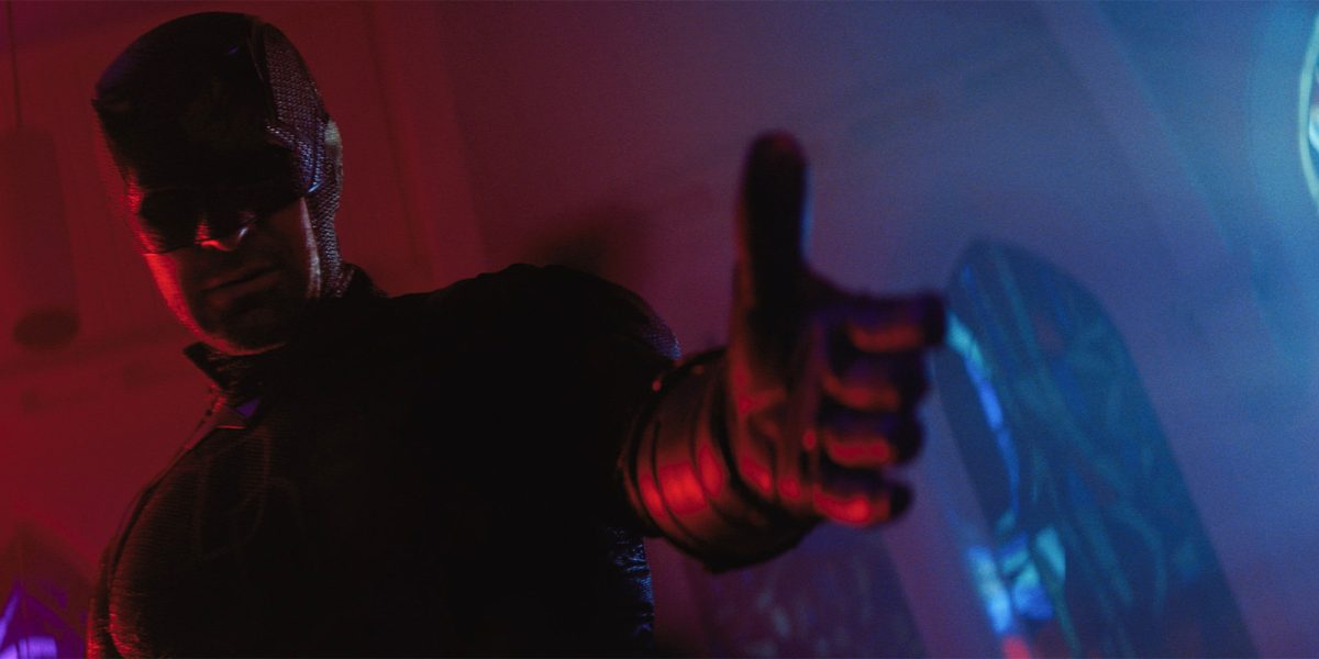 A person in a dark, armoured costume with a mask is standing indoors, extending their right arm forward with their hand in a gesture resembling a gun. The background features stained glass windows with blue and red lighting.