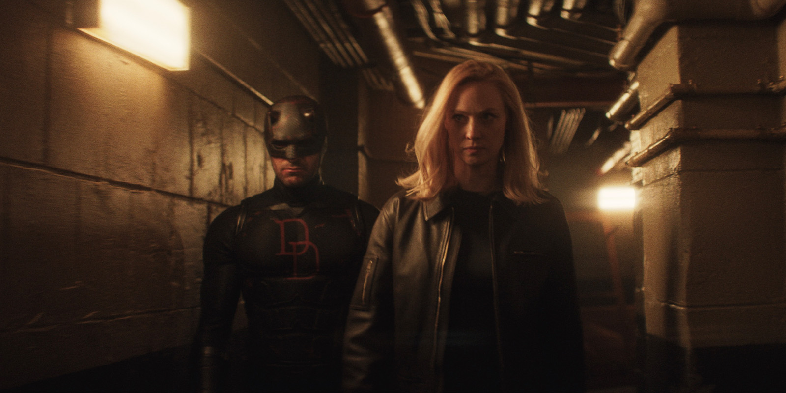 A man in a black superhero costume with a red DD on his chest walks behind a blonde woman in a black jacket down a dimly lit industrial corridor with exposed pipes on the ceiling.