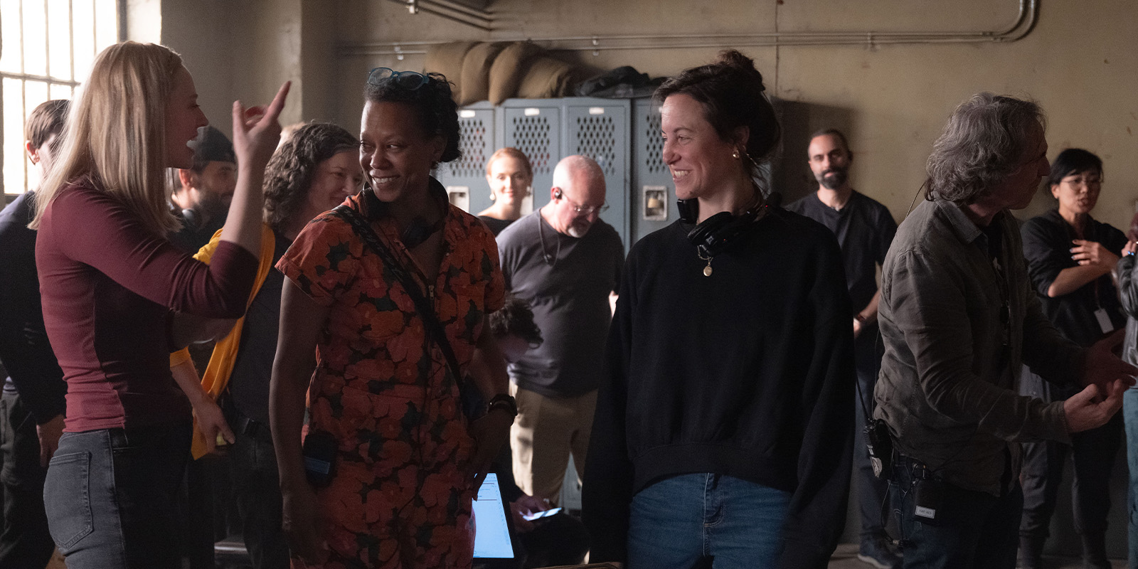 A group of people stand and talk in a room with lockers on the back wall. Some are smiling and engaged in conversation, whilst others appear to be listening or interacting in small groups. The lighting is soft and natural.