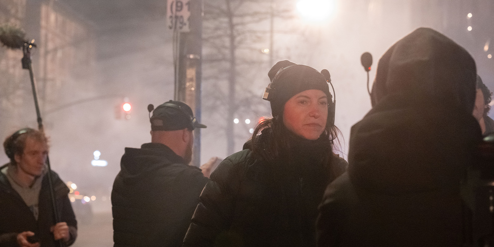 Several people wearing headsets and winter clothing stand on a city street at night, surrounded by mist or smoke. One person holds a boom microphone. A street sign and traffic lights are visible in the background.