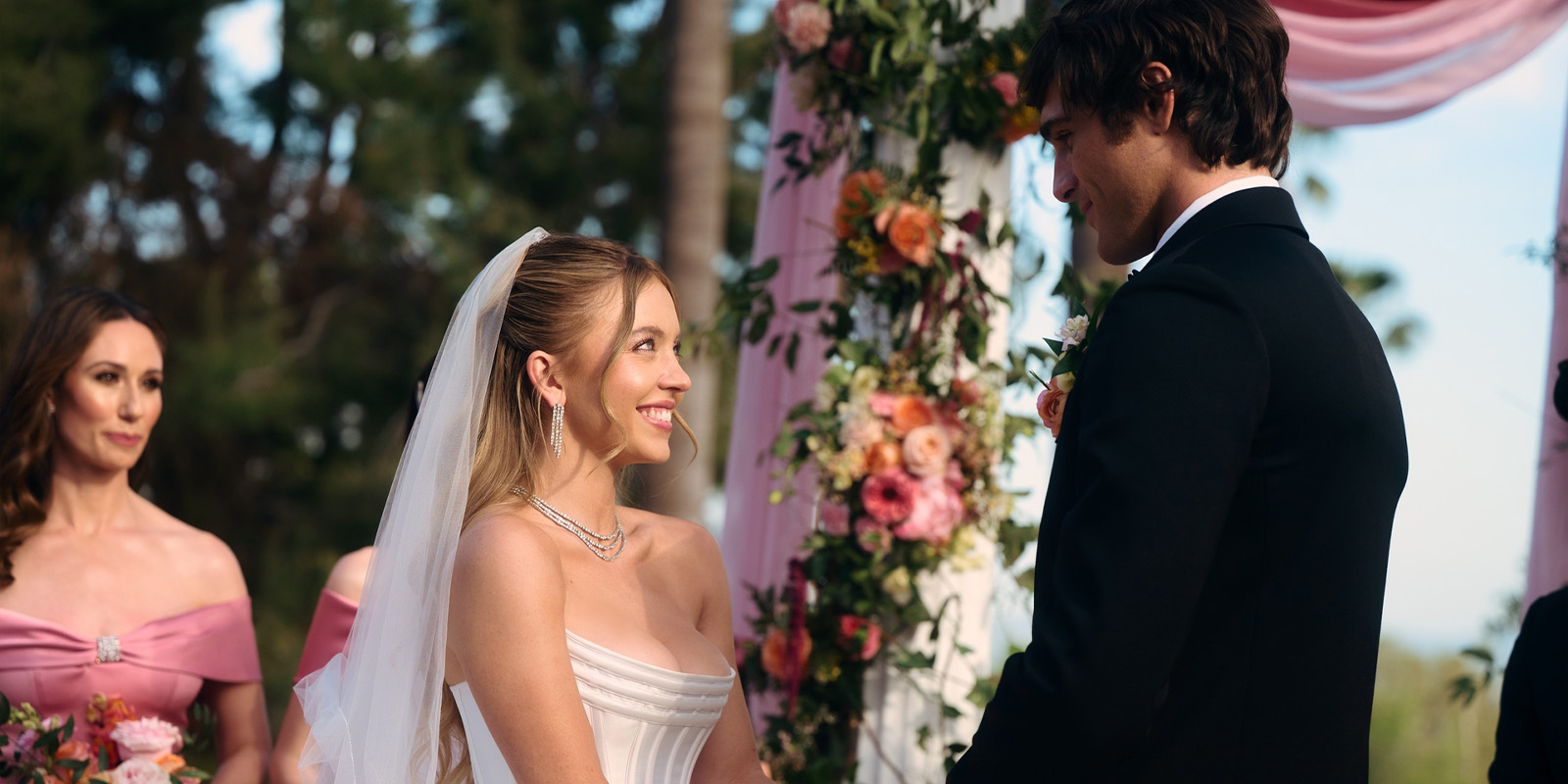 A bride and groom stand facing each other, holding hands and smiling, during an outdoor wedding ceremony with floral decorations. A bridesmaid in a pink dress stands to the side, watching the couple.