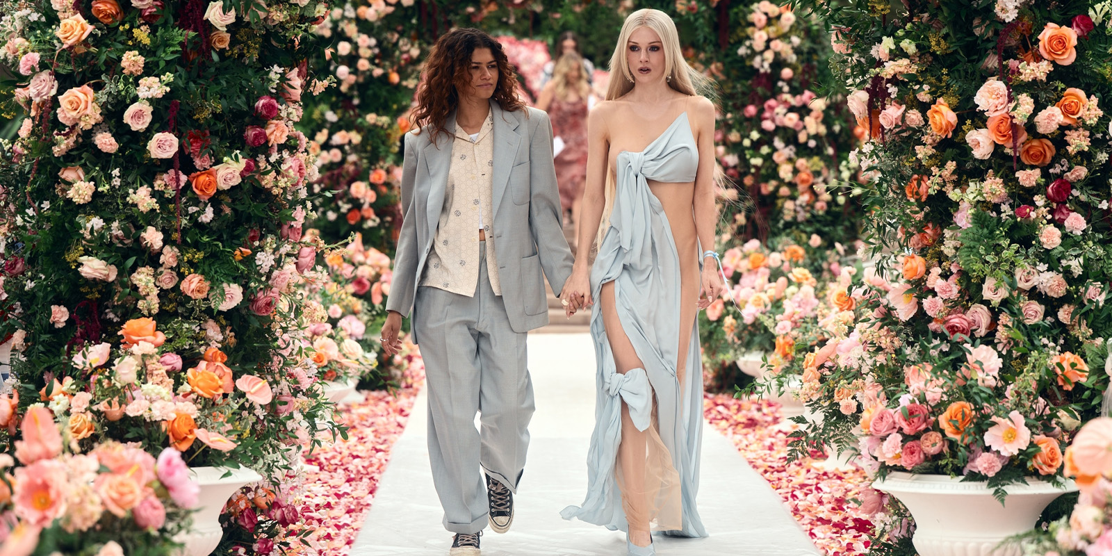 Two people walk hand-in-hand down a flower-lined aisle. One wears a light grey suit and trainers, the other wears a flowing pale blue dress with a high slit. Both have long hair, and lush floral arches surround them.