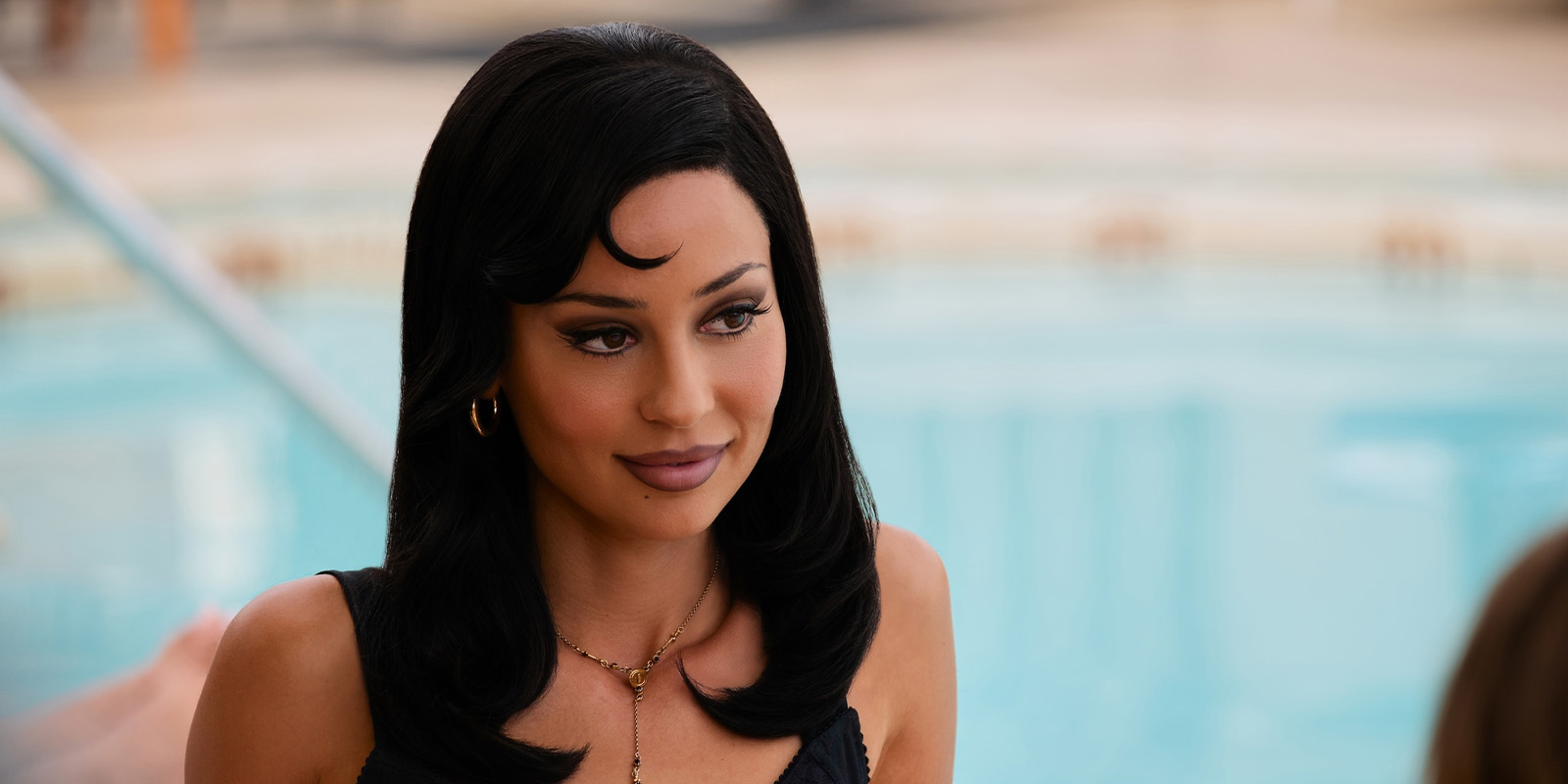 A woman with long dark hair and gold jewellery sits by a swimming pool, wearing a dark top and looking slightly to the side with a neutral expression.