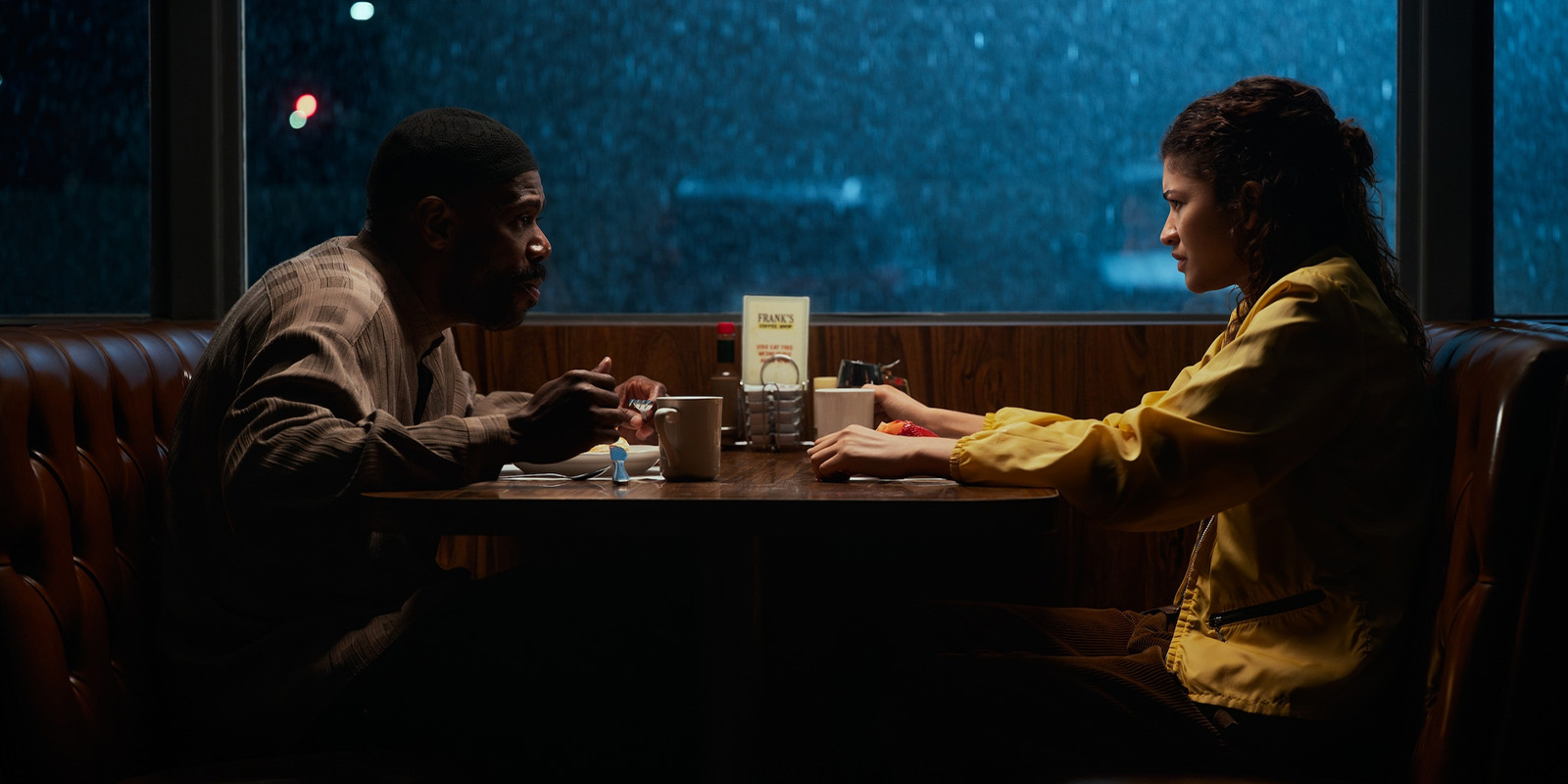 A man and a woman sit opposite each other at a booth in a dimly lit diner at night, both with serious expressions, mugs and plates of food on the table, and rain visible through the window behind them.