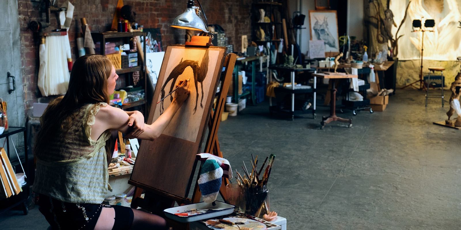 A person sits at an easel in an art studio, painting a picture of a tree. Art supplies, brushes, and paintings are visible on tables and shelves around the spacious studio with brick walls.