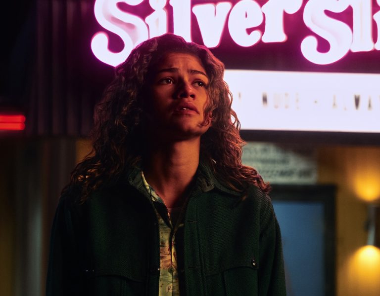 A young woman with curly hair stands outside at night, illuminated by a neon sign reading Silver Slipper. She wears a dark jacket and looks slightly upward with a serious expression.