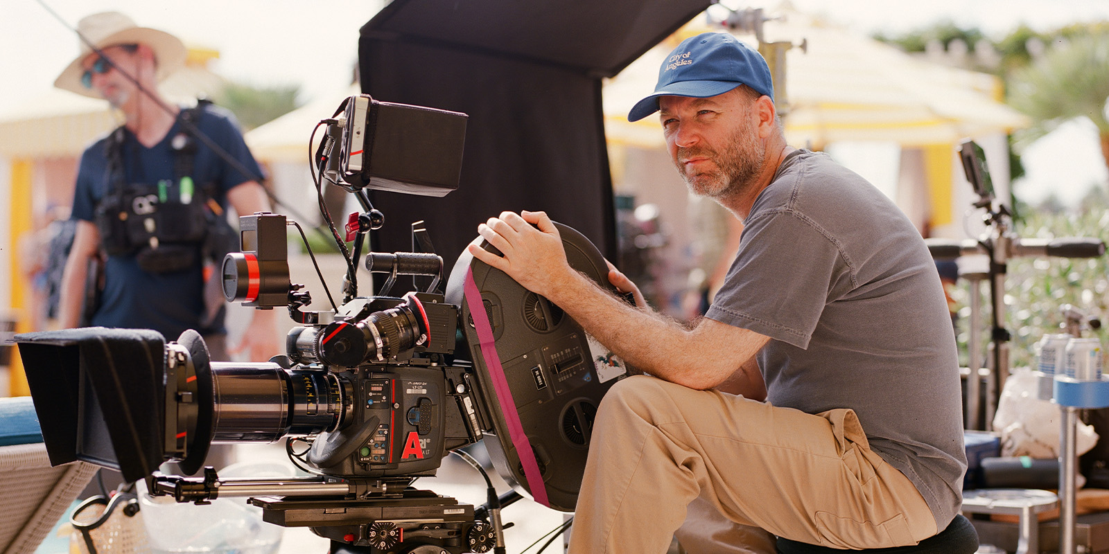 A man wearing a blue cap and beige trousers sits at a film camera on an outdoor set, holding the camera's controls. Another person and filming equipment are visible in the background.