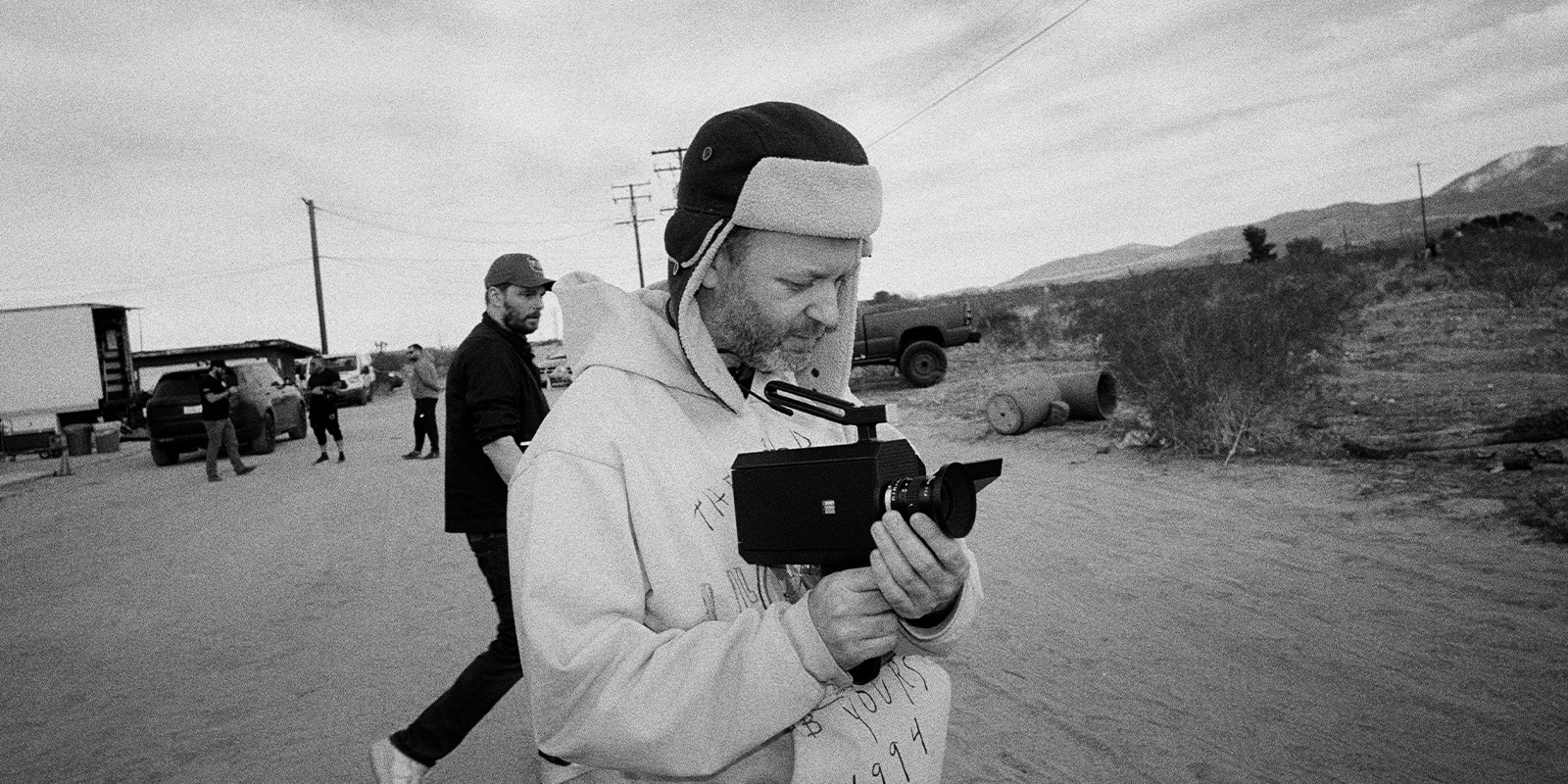 A person wearing a hoodie and a cap is holding a vintage video camera on a dirt road, with another person walking behind and vehicles parked in the background under a cloudy sky.