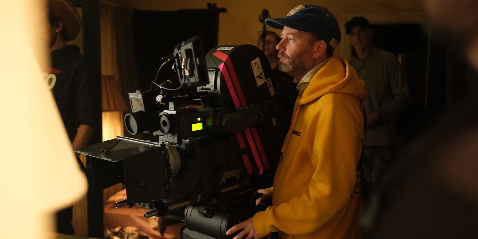 A person in a yellow hoodie and blue cap operates a large professional film camera on set, with crew members and filming equipment visible in the background.