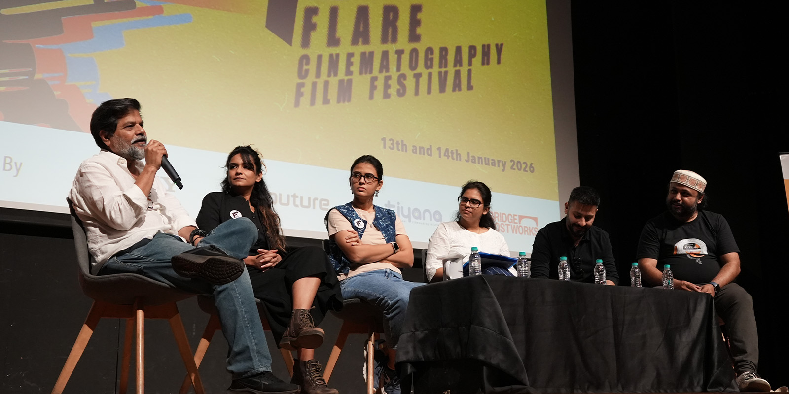 Six panellists are seated on stage during a discussion at the Flare Cinematography Film Festival, with one man speaking into a microphone. A large event banner is visible behind them, showing the festival dates.
