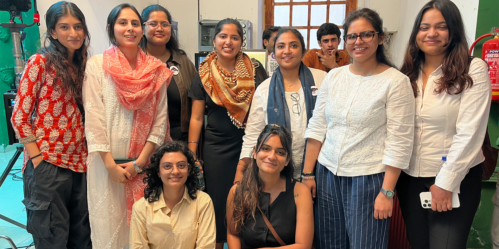 A group of ten people, mostly women, pose together indoors. Most are standing, a few are seated in front. They are casually dressed and smiling at the camera, with laboratory equipment visible in the background.