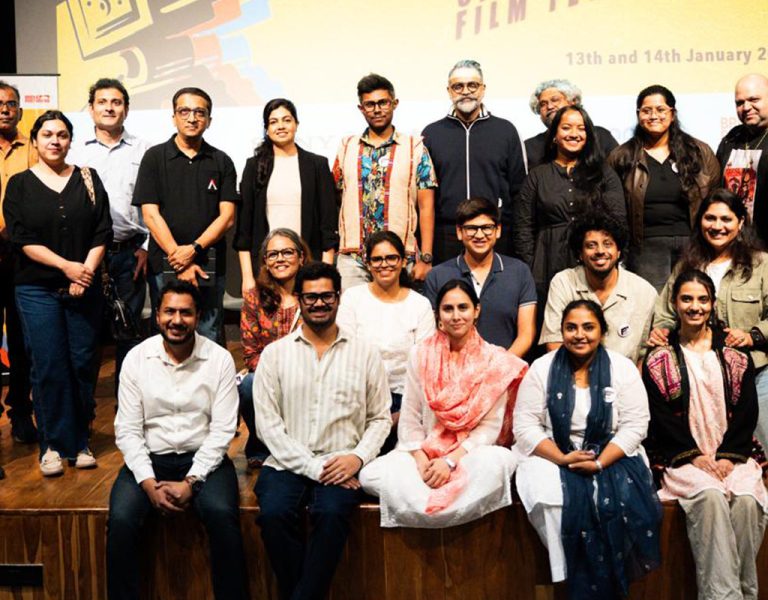 A group of people stand and sit together on a wooden stage for a group photo. They are facing the camera and smiling. A presentation screen with colourful graphics and text is in the background.