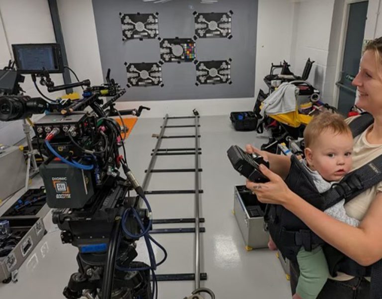 A woman holds a baby in a front carrier while operating camera equipment in a room with a track, toolboxes, and technical gear. The baby looks forward while the woman smiles and focuses on the camera.