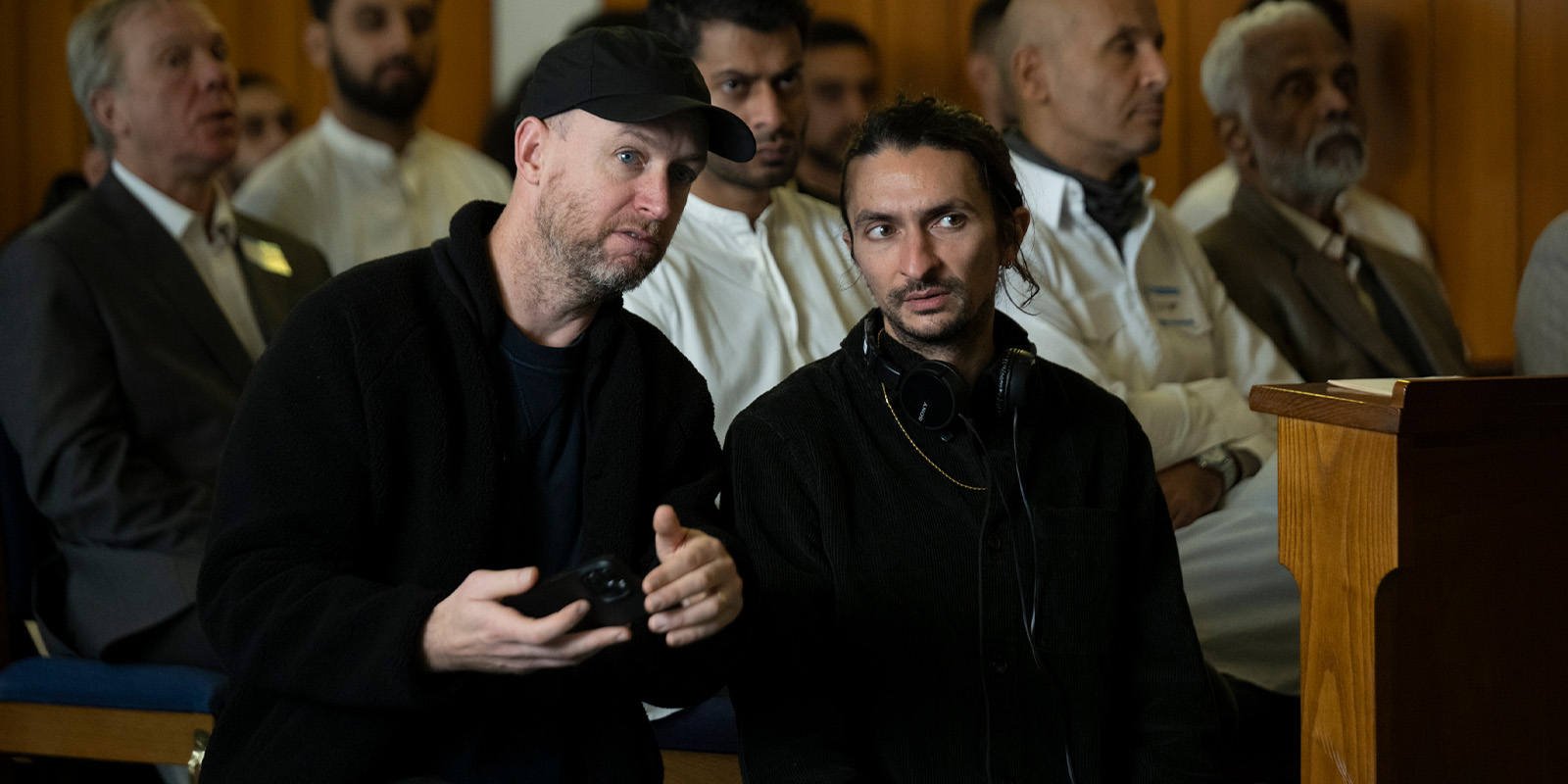 Two men sit in the foreground of a room, one holding a device and the other wearing headphones around his neck, with several people seated in the background, all facing forwards.