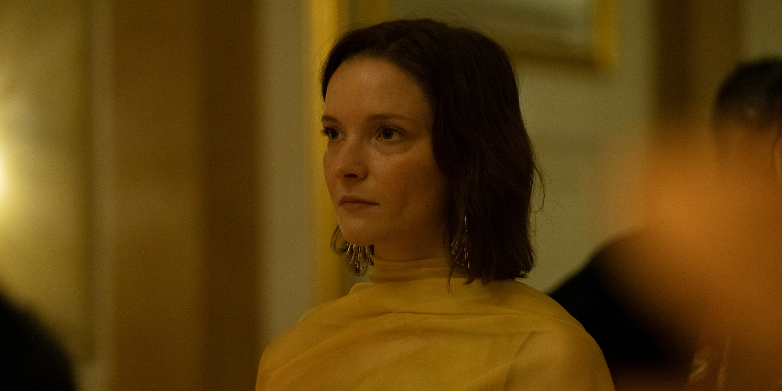 A woman with shoulder-length brown hair sits indoors, wearing a sheer yellow top and dangling earrings. She looks slightly to the side with a neutral expression. The background is softly lit and out of focus.
