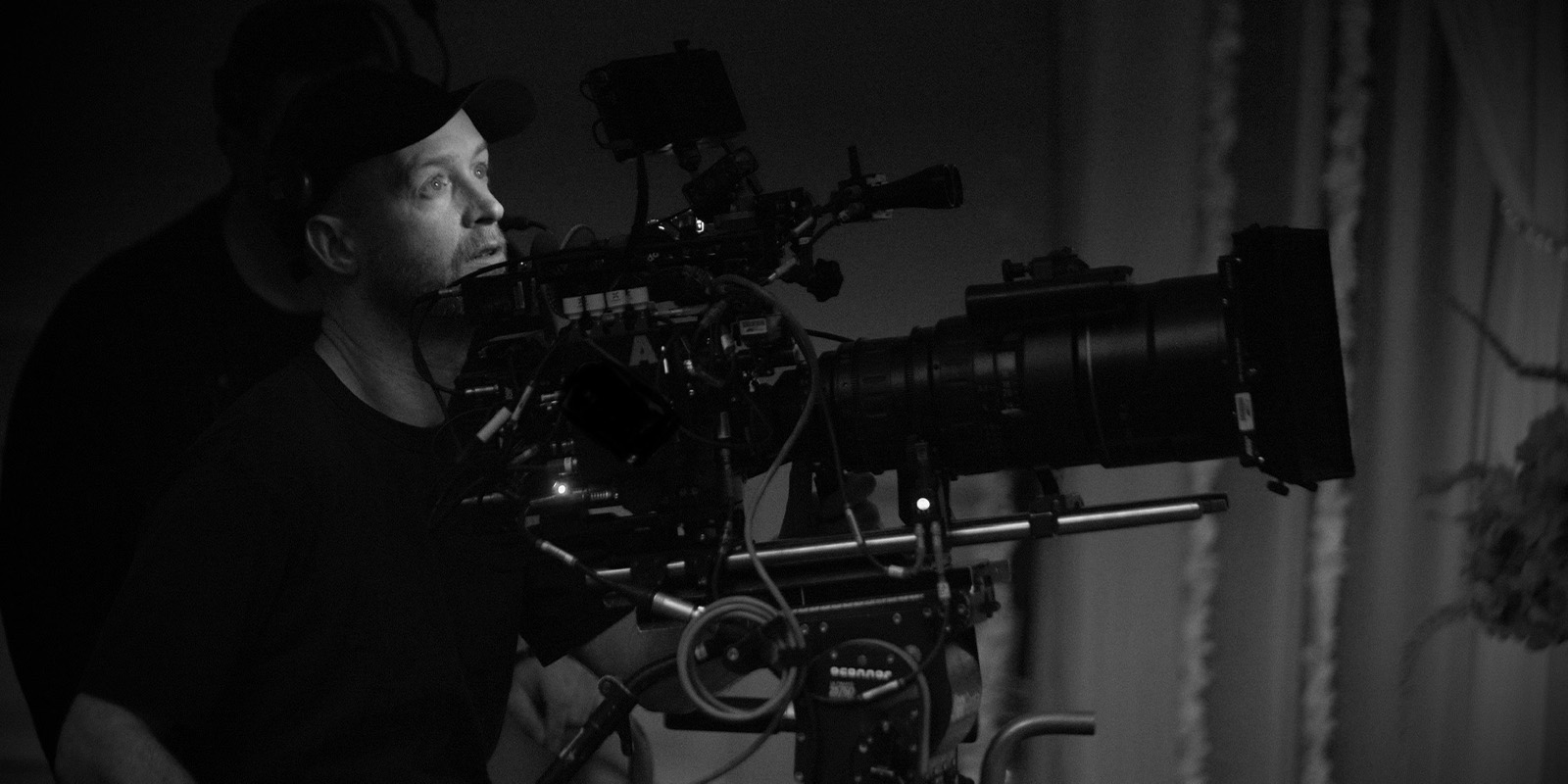 A person wearing a cap operates a large professional video camera mounted on a tripod, focusing intently on the scene. The surroundings are dimly lit and the image is in black and white.