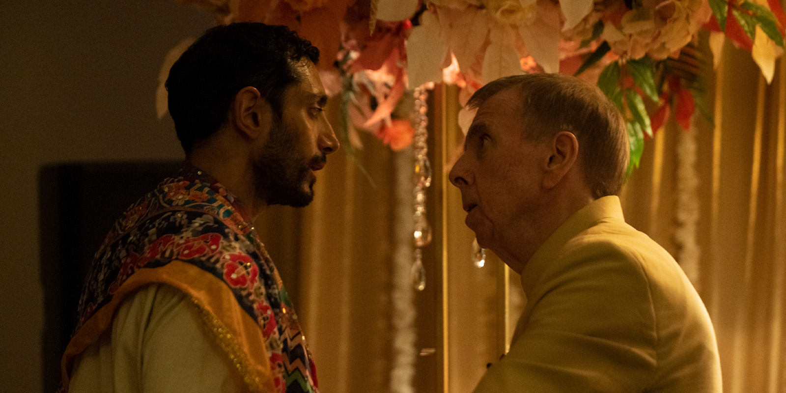 Two men stand close together in conversation under a floral arrangement. One man wears a colourful patterned garment, while the other wears a light yellow suit. Warm lighting and curtains create a soft background.