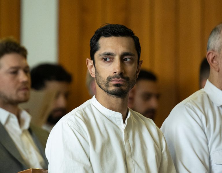 A group of men, most wearing white shirts, stand in rows indoors. The man in the centre foreground has short dark hair and is looking to his left. The background shows wood-panelled walls.